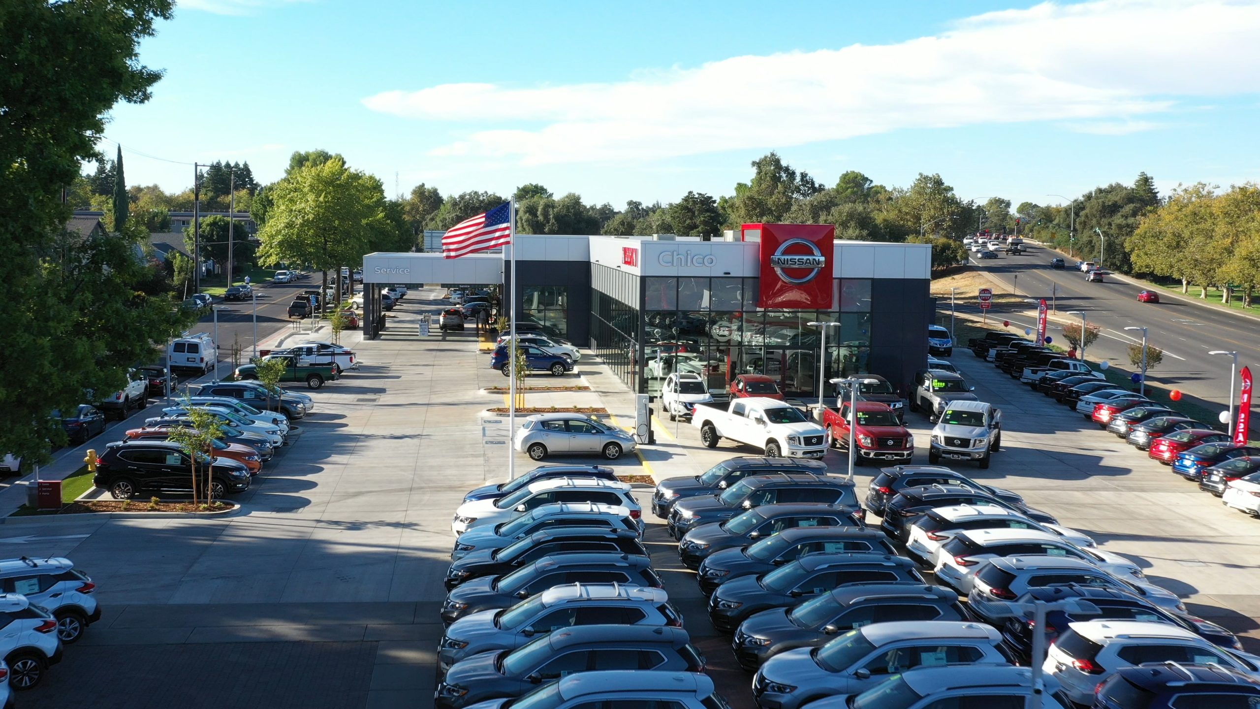 Aerial view of Chico Nissan dealership with rows of new and used vehicles, showroom building, service drive, and American flag in Chico, California.